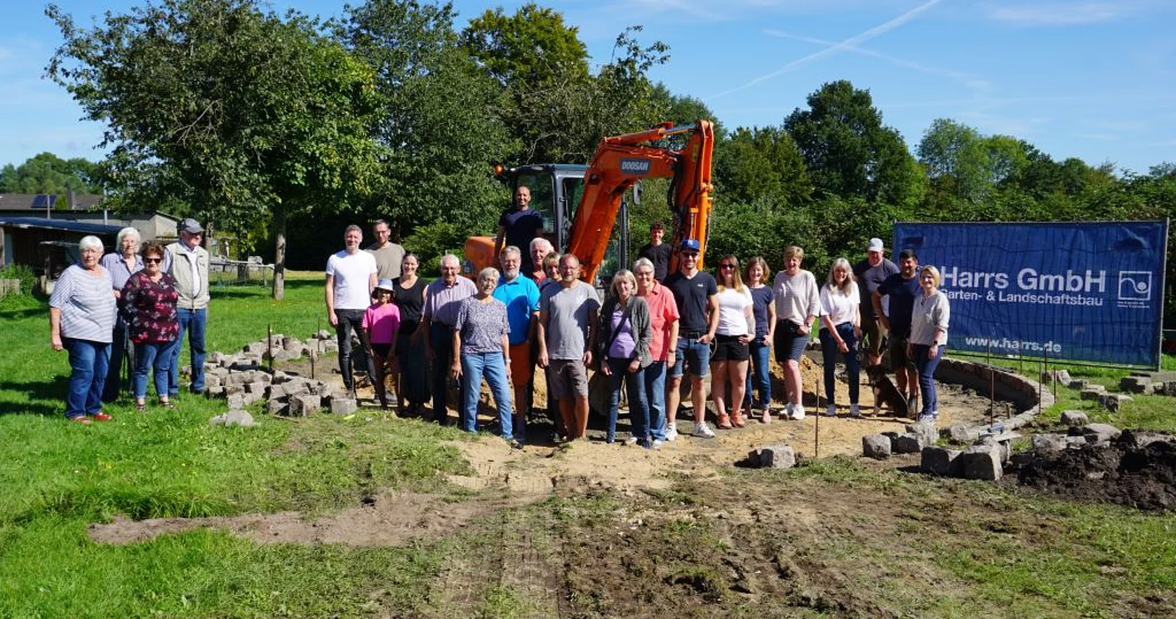 Eine Gruppe von Menschen steht an der Baustelle und schaut in die Kamera. Ein Bagger ist im Hintergrund zu sehen.