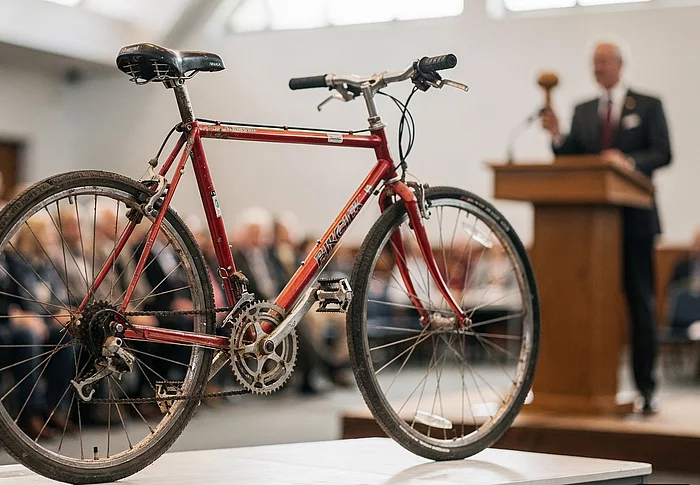 Ein gebrauchtes Fahrrad steht auf einem Tisch. Im Hintergrund ist verschwommen ein Mann zu sehen der einen Hammer in der Hand hält.