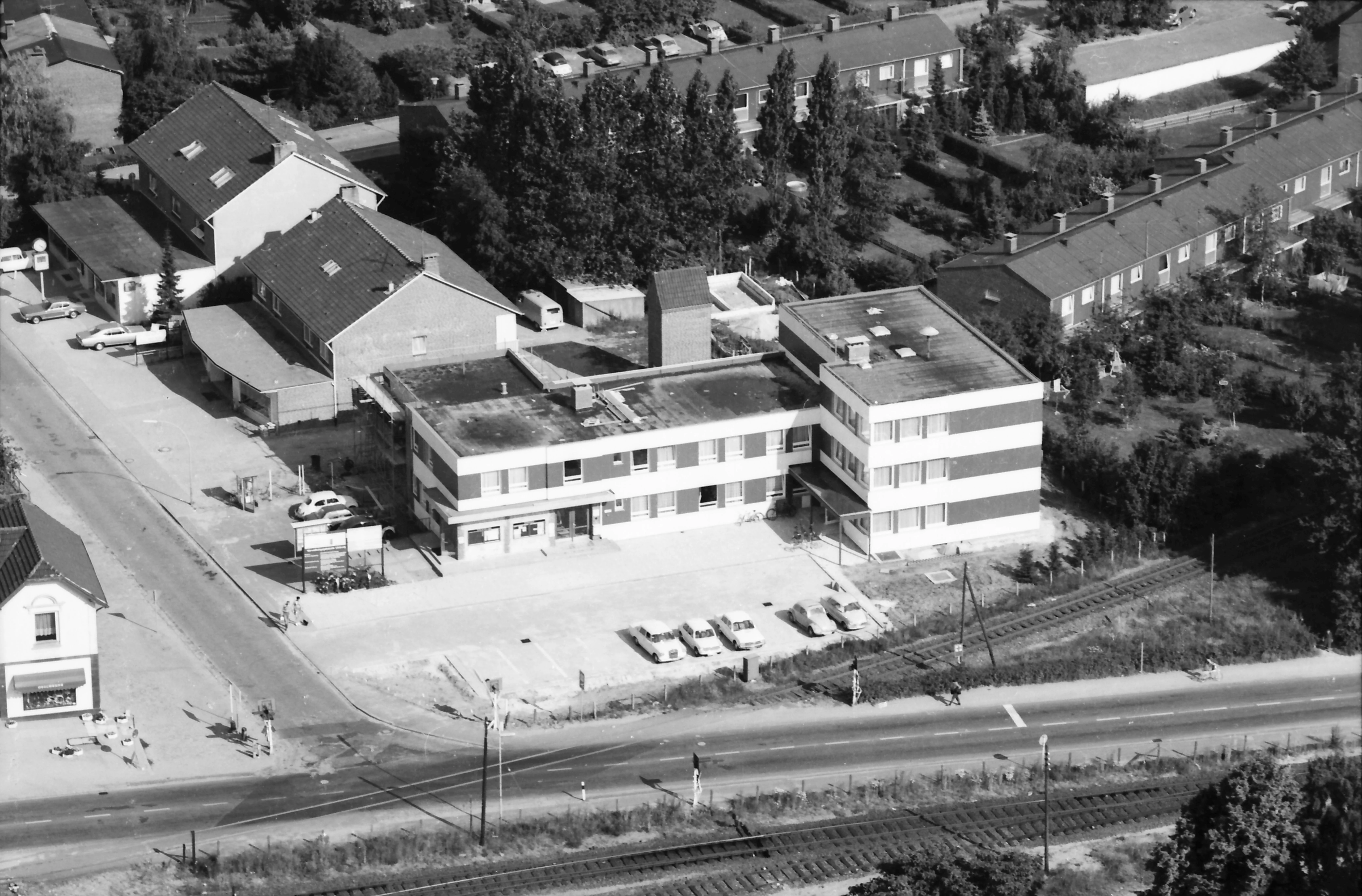 Zu sehen ist eine Luftbildaufnahme in Schwarz-Weiß. Zentral in der Bildmitte ist das alte Verwaltungsgebäude der Gemeinde Henstedt-Ulzburg circa Anfang der 1970er Jahre zu sehen, welches sich an der Ecke Hamburger Straße/Beckersbergstraße befand. 