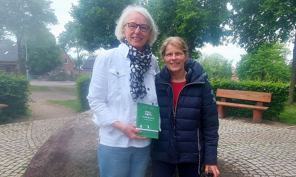 Zwei Frauen posieren für das Foto und halten ein Buch in der Hand. Im Hintergrund ist ein Park zu sehen.