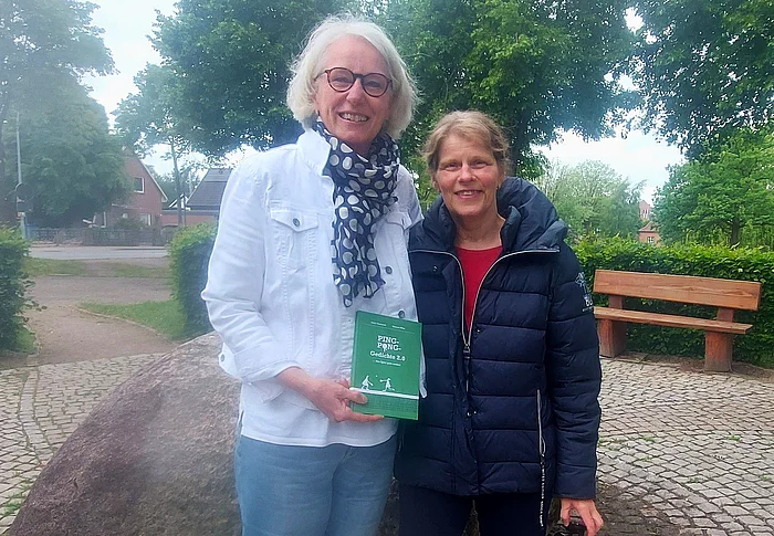 Zwei Frauen posieren für das Foto und halten ein Buch in der Hand. Im Hintergrund ist ein Park zu sehen.
