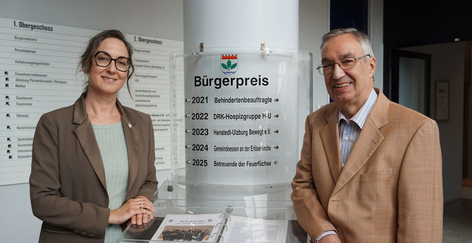 Bürgermeisterin Ulrike Schmidt und Bürgervorsteher Henry Danielski stehen vor der Säule mit den bisherigen Bürgerpreisträgerinnen und -trägern