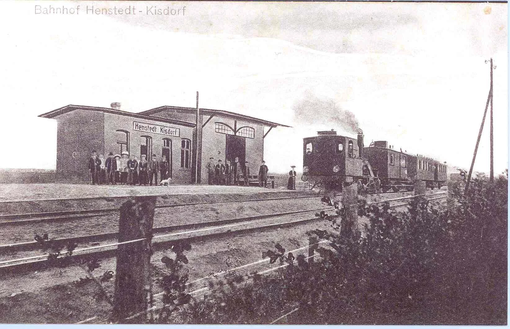 Das Bild zeigt eine Schwarz-Weiß-Aufnahme des Bahnhofs Henstedt – Kisdorf. Vor dem Gebäude befindet sich eine zweigleisige Bahntrasse. In der rechten Bildhälfte steht auf dem hinteren Gleis eine kleine Dampflokomotive mit zwei Waggons. Vor dem Bahnhofsgebäude stehen zwei gemischte Gruppen von Personen. 