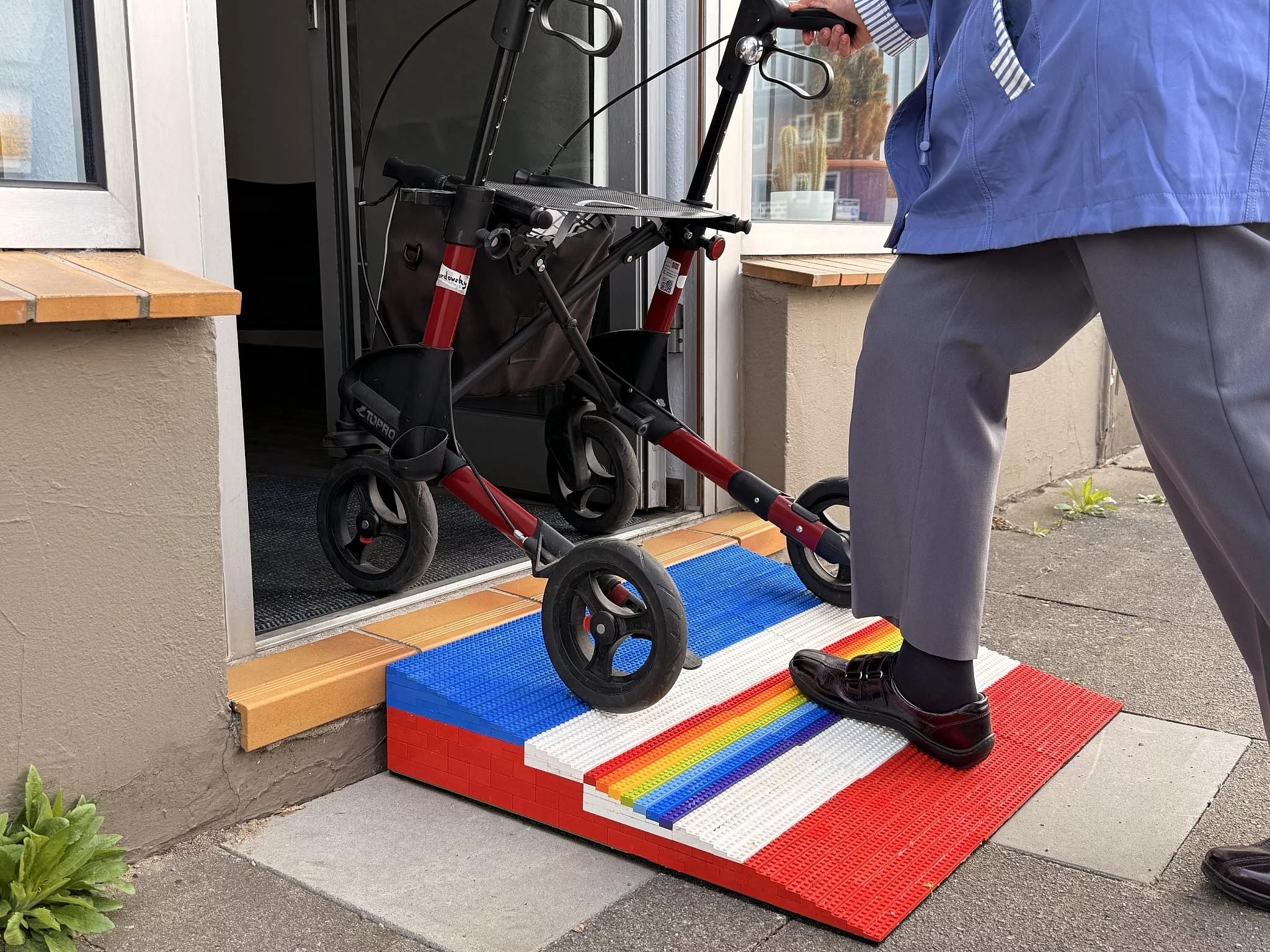 Eine Person schiebt einen Rollator über eine bunte Lego-Rampe, die eine Stufe vor einer Hauseingangstür überbrückt.