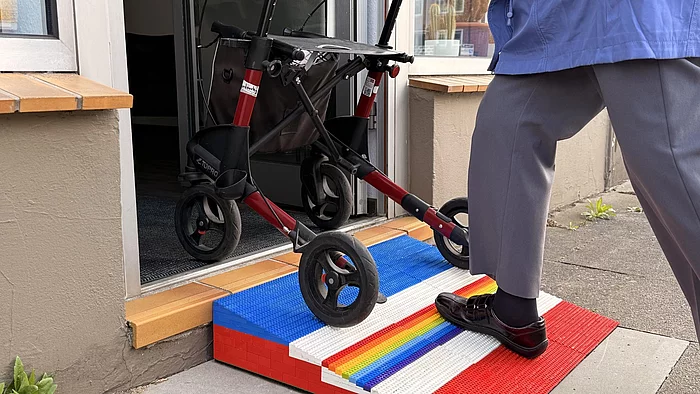 Eine Person schiebt einen Rollator über eine bunte Lego-Rampe, die eine Stufe vor einer Hauseingangstür überbrückt.