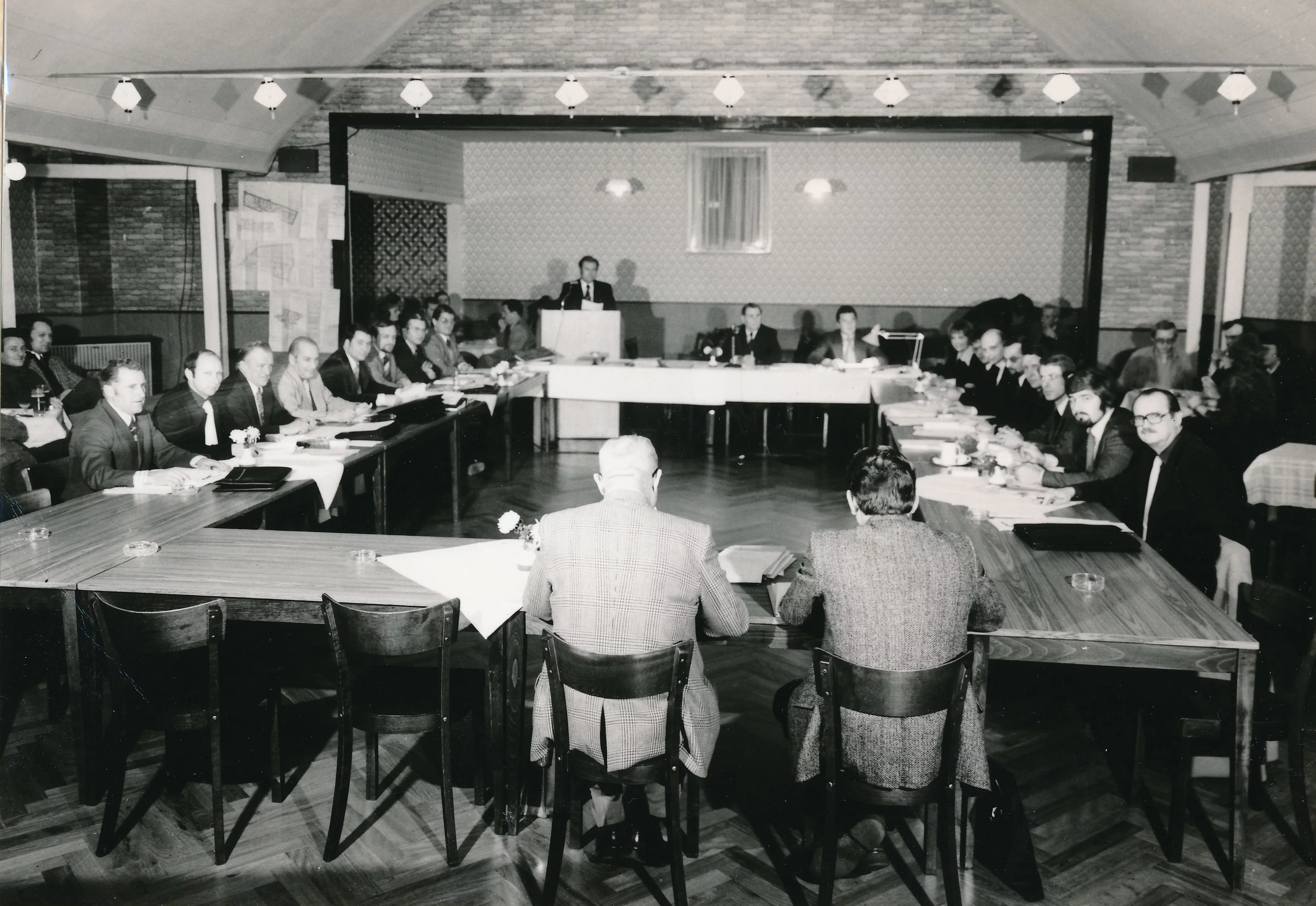 Abgebildet ist die Gemeindevertretung der Wahlperiode 1974 bis 1978 bei einer Sitzung im Gasthof Scheelke. Es ist ein Schwarz-Weiß-Foto. Im Bildhintergrund steht Bürgermeister Glück an einem Rednerpult. Die übrigen Vertreterinnen und Vertreter der Gemeinde, die mehrheitlich Männer sind, sitzen an Tischen. 