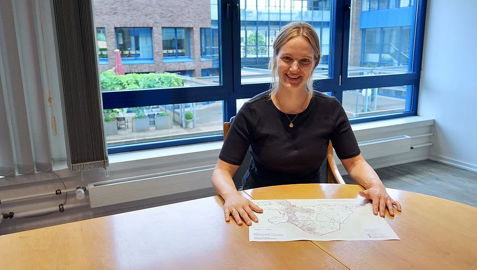 Klimaschutzmanagerin Hannah Grünewald sitz an einem Tisch im Rathaus und lächelt in die Kamera. Vor ihr liegt der Plan zur kommunalen Wärmeplanung.