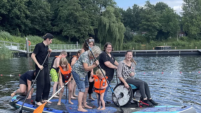 Eine Gruppe von Erwachsenen und Kindern, darunter eine Frau im Rollstuhl, steht und sitzt gemeinsam auf einem riesigen Stand-up-Paddle-Board (SUP) auf einem See.